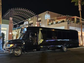 Black party bus parked at night in front of a lit outdoor shopping and dining plaza with string lights, rooftop patio seating, palm trees and holiday lights.