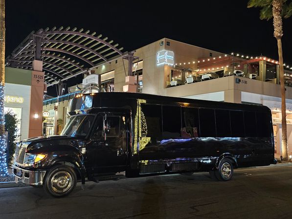 Black party bus parked at night in front of a lit outdoor shopping and dining plaza with string lights, rooftop patio seating, palm trees and holiday lights.