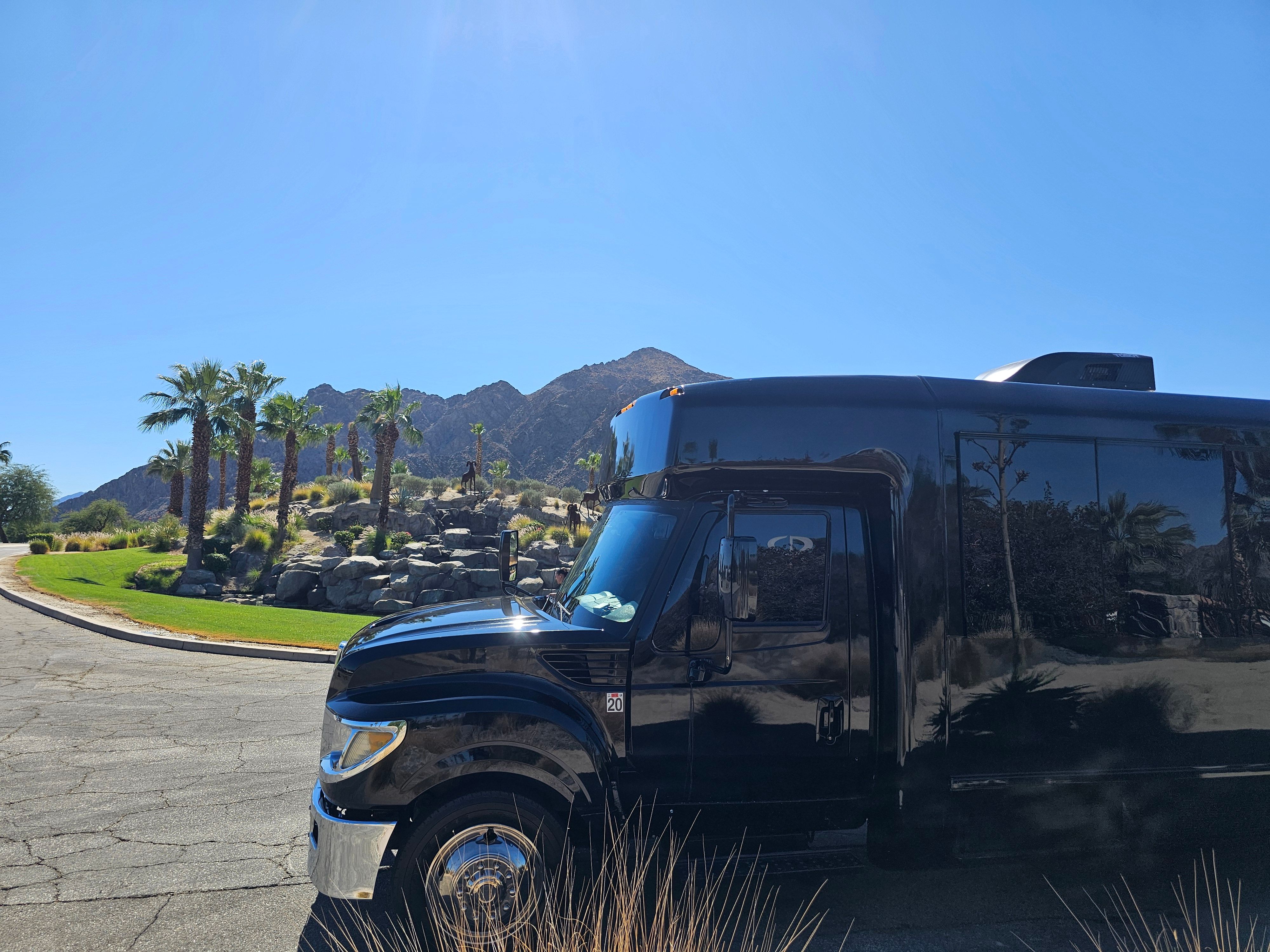 Sleek black shuttle bus parked beside a palm‑lined desert rock garden and rugged mountains under a clear blue sky