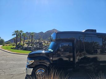 Sleek black shuttle bus parked beside a palm‑lined desert rock garden and rugged mountains under a clear blue sky