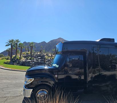 Sleek black shuttle bus parked beside a palm‑lined desert rock garden and rugged mountains under a clear blue sky