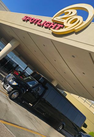 Black luxury shuttle/party bus parked under a beige event venue entrance canopy with a large circular marquee sign, glass doors and a clear blue sky overhead.