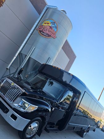 Black passenger shuttle bus parked beside a tall corrugated metal brewery silo decorated with a colorful sun-and-palm emblem, photographed at an angle against a clear blue sky.