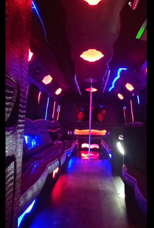 Neon-lit party bus interior with a center dance pole, wraparound leather bench seating and vibrant pink, blue and purple LED ambient lights.