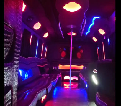 Neon-lit party bus interior with a center dance pole, wraparound leather bench seating and vibrant pink, blue and purple LED ambient lights.
