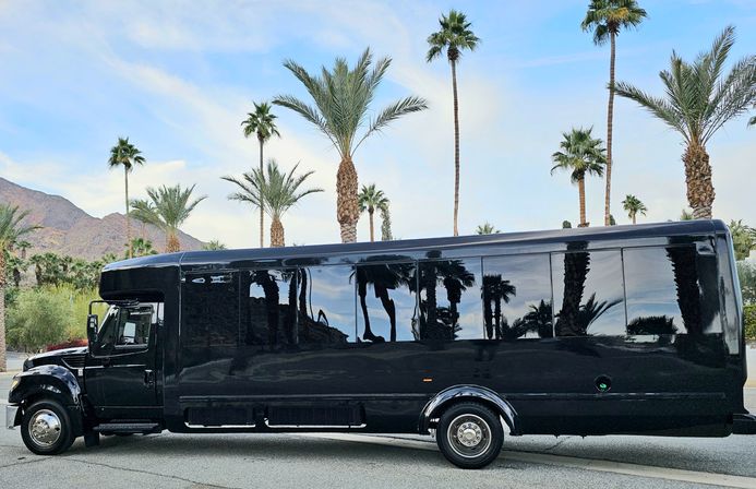 Black party bus parked on a palm-lined California desert street, glossy windows reflecting tall palm trees and distant mountains beneath a blue sky.
