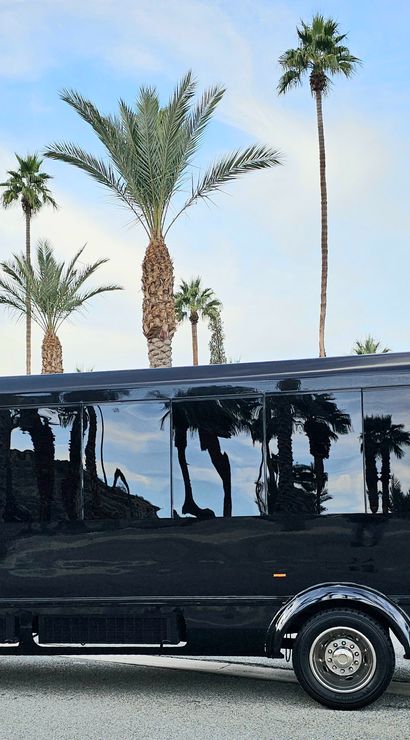 Black party bus parked on a palm-lined California desert street, glossy windows reflecting tall palm trees and distant mountains beneath a blue sky.