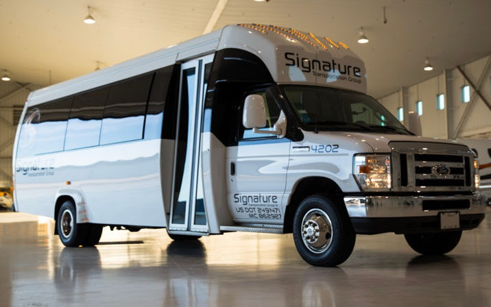 White passenger shuttle minibus with open folding doors parked inside a bright aircraft hangar, chrome grille and tinted windows reflecting on the polished concrete floor.