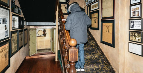 Visitor in a parka climbing a carpeted staircase with a wooden banister in a small history museum hallway, walls lined with framed vintage newspapers and photos and an open safe under the stairs.