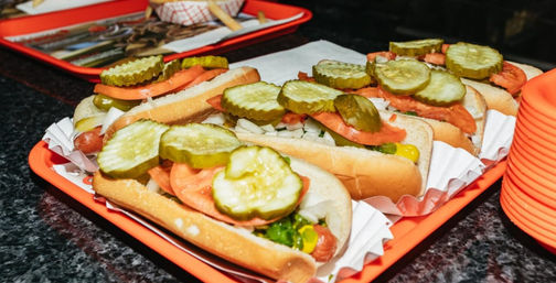 Tray of Chicago-style hot dogs on orange trays topped with large dill pickle slices, tomato wedges, chopped onions, mustard and bright green relish — casual fast-food scene