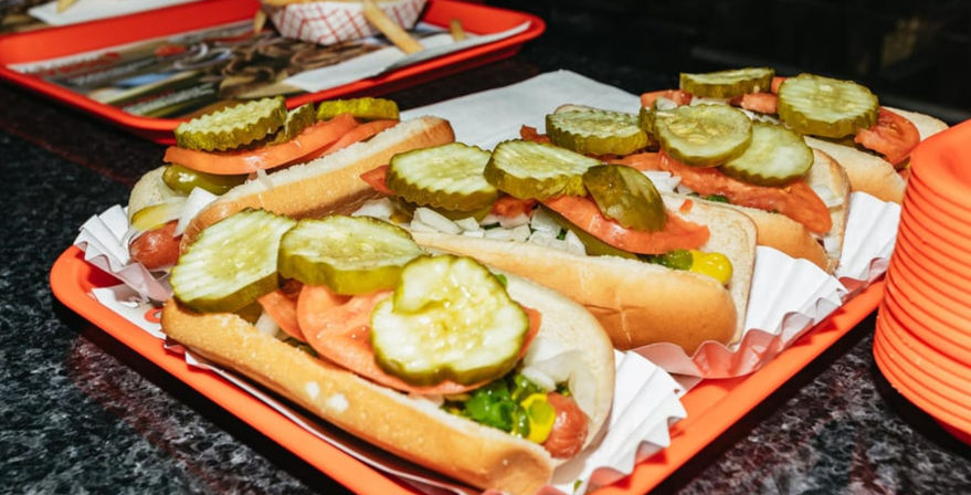 Tray of Chicago-style hot dogs on orange trays topped with large dill pickle slices, tomato wedges, chopped onions, mustard and bright green relish — casual fast-food scene