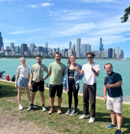 Six friends posing on a grassy Lake Michigan shoreline with the Chicago skyline and blue sky in the background.