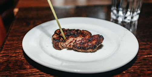 Charred grilled steak medallion pierced by a bamboo skewer on a white plate set on a dark wooden table with a water glass blurred in the background.