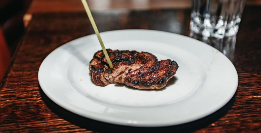 Charred grilled steak medallion pierced by a bamboo skewer on a white plate set on a dark wooden table with a water glass blurred in the background.