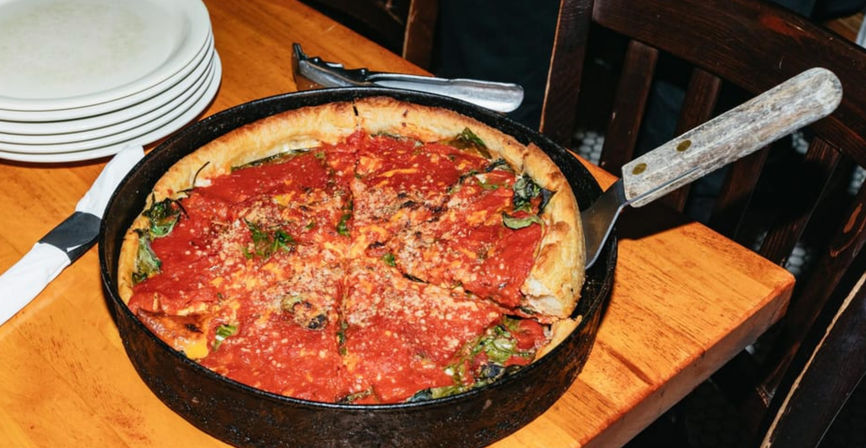 Skillet deep-dish pizza topped with chunky tomato sauce, melted cheese and greens, served in a cast-iron pan with a spatula on a wooden restaurant table beside stacked plates.