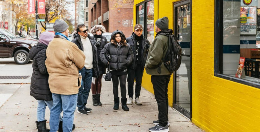 Small group in winter jackets gathered on a city sidewalk beside a bright yellow storefront and brick buildings, listening to a person speaking.