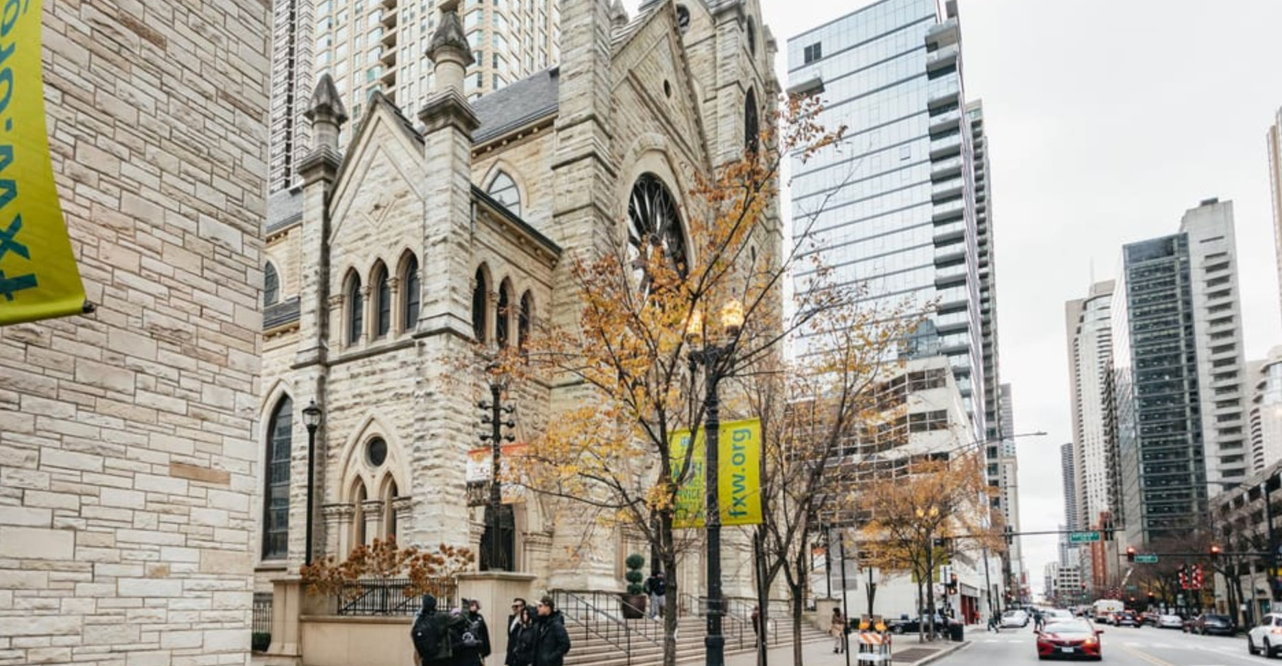 Historic stone church beside glass skyscrapers on a downtown city street in autumn, yellow-leaved trees, pedestrians and cars passing under green banners.