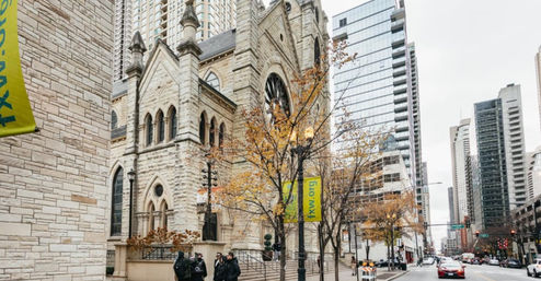 Historic stone church beside glass skyscrapers on a downtown city street in autumn, yellow-leaved trees, pedestrians and cars passing under green banners.