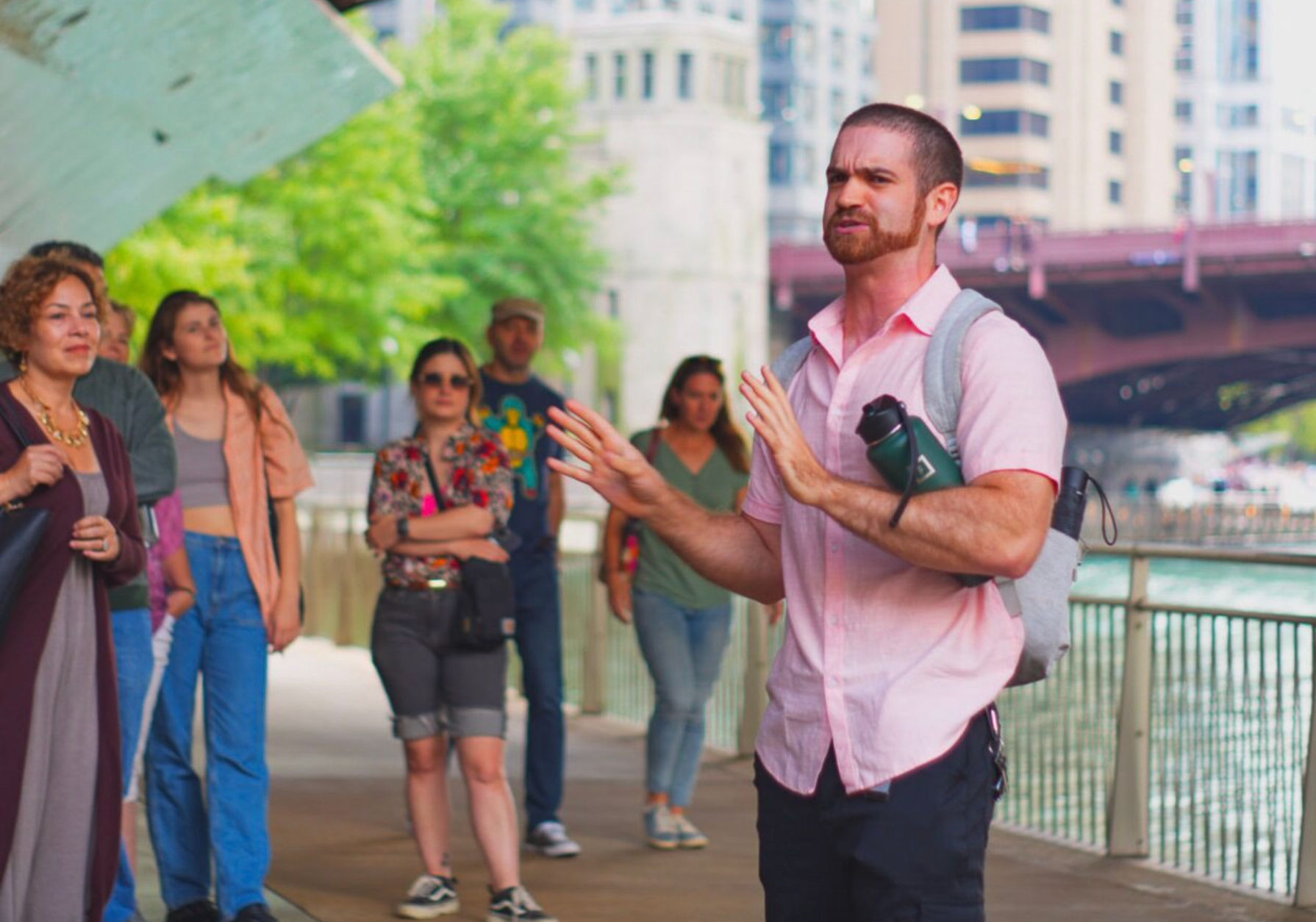 Tour guide animatedly addressing a small group on a city riverfront walkway under a bridge, with water and skyline in the background.