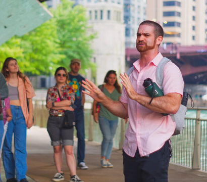 Tour guide animatedly addressing a small group on a city riverfront walkway under a bridge, with water and skyline in the background.