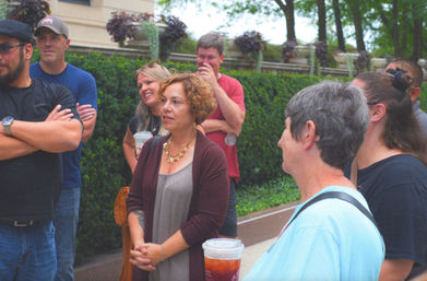 Outdoor city walking tour group gathered on a sidewalk by manicured hedges, adults chatting and holding iced drinks.
