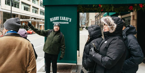 Tour guide gestures on a downtown city sidewalk beneath a green restaurant awning while a small group of bundled-up pedestrians in winter coats and fur-lined hoods listens.