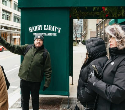 Tour guide gestures on a downtown city sidewalk beneath a green restaurant awning while a small group of bundled-up pedestrians in winter coats and fur-lined hoods listens.