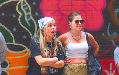 Two young women in summer outfits—one with a bandana and surprised expression, the other in sunglasses and a white crop top—standing in front of a colorful urban graffiti mural on a city wall