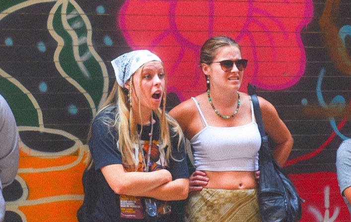 Two young women in summer outfits—one with a bandana and surprised expression, the other in sunglasses and a white crop top—standing in front of a colorful urban graffiti mural on a city wall