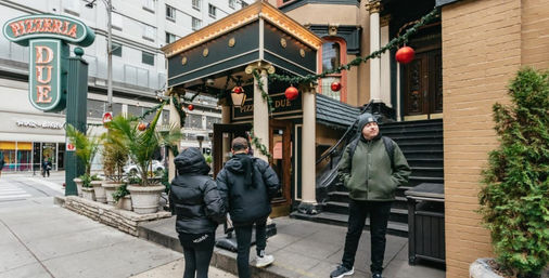 Urban downtown sidewalk scene with people in winter coats outside a decorated pizzeria entrance featuring garlands, red holiday ornaments, large planters and stairs, with city buildings in the background.