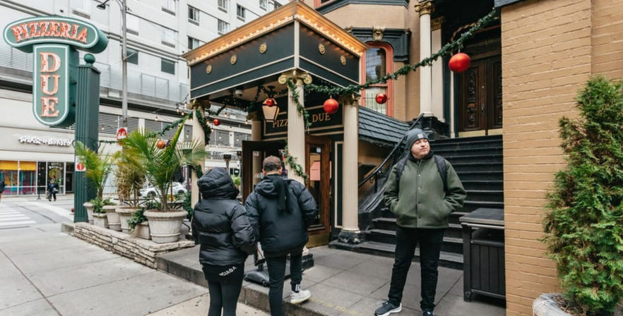 Urban downtown sidewalk scene with people in winter coats outside a decorated pizzeria entrance featuring garlands, red holiday ornaments, large planters and stairs, with city buildings in the background.