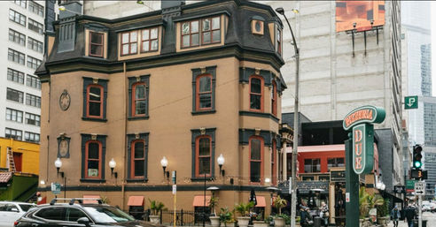 Charming historic three-story brownstone with mansard roof on a downtown street corner, ground-floor pizzeria/restaurant with outdoor seating, string lights, street lamps, parked cars and high-rise buildings in the background.