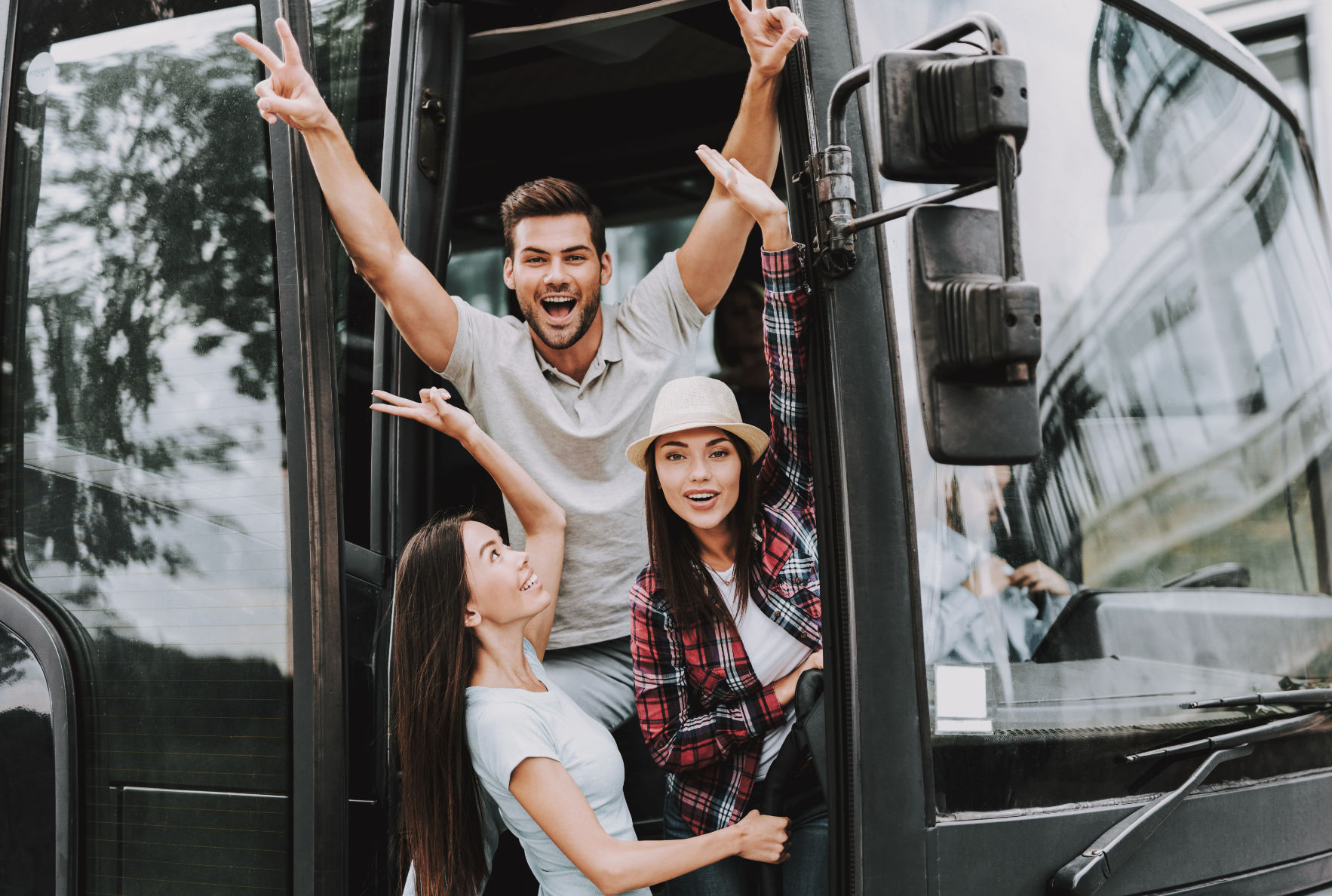 Three cheerful friends waving from the open door of a tour bus on an urban group trip