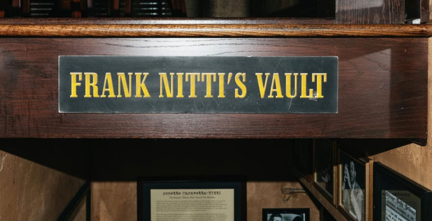 Wood-paneled stairway entrance to a speakeasy-style vault with a black plaque in bold yellow letters and framed photos and historical documents lining the walls.