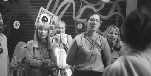Black-and-white candid street photo of four women giving surprised and curious looks, with another person seen from behind, standing in front of a graffiti-covered metal shutter in a downtown urban scene.