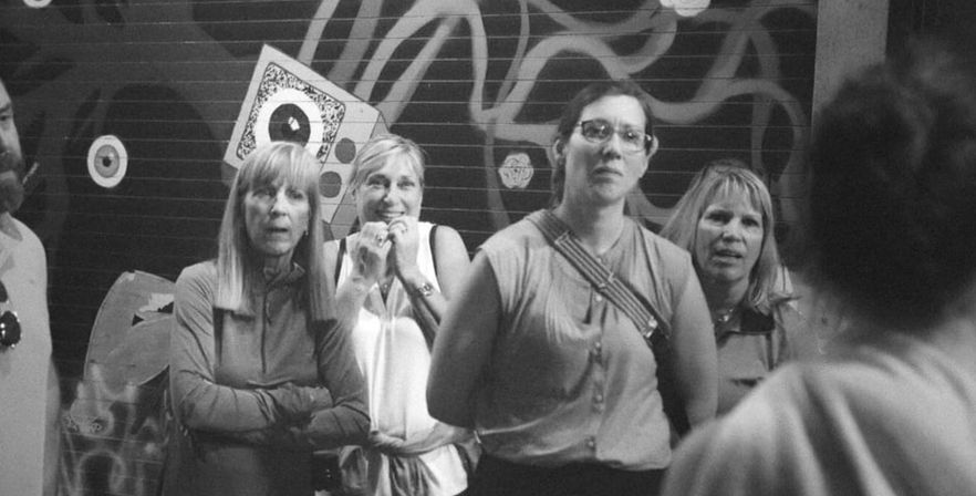 Black-and-white candid street photo of four women giving surprised and curious looks, with another person seen from behind, standing in front of a graffiti-covered metal shutter in a downtown urban scene.