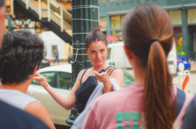 Animated woman gesturing while chatting with two friends on a busy city sidewalk, parked cars and a metal staircase visible in the background.