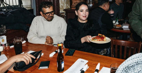 People at a cozy, bustling pizzeria seated around a wooden table; one person holds a plate with a deep-dish pizza slice amid drinks, menus, and napkins.