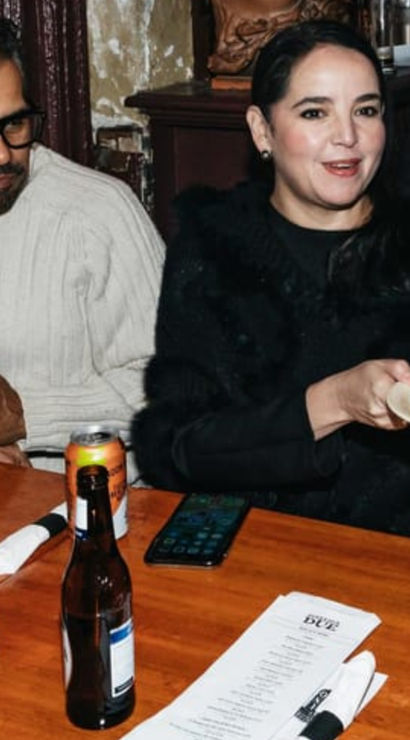 People at a cozy, bustling pizzeria seated around a wooden table; one person holds a plate with a deep-dish pizza slice amid drinks, menus, and napkins.