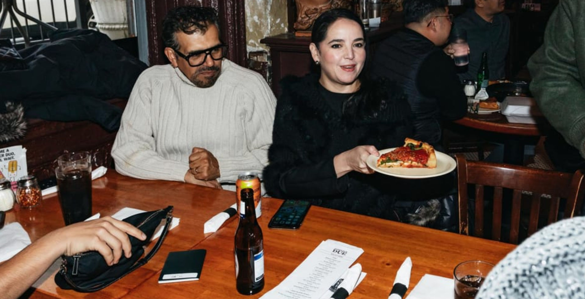 People at a cozy, bustling pizzeria seated around a wooden table; one person holds a plate with a deep-dish pizza slice amid drinks, menus, and napkins.