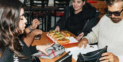 Three people at a casual diner table passing a red tray of loaded hot dogs topped with pickles, tomato slices and onions — friends sharing a quick bite.