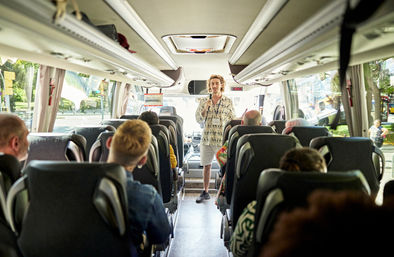 Cheerful tour guide standing and addressing seated passengers inside a modern coach bus on a sunny city sightseeing tour.