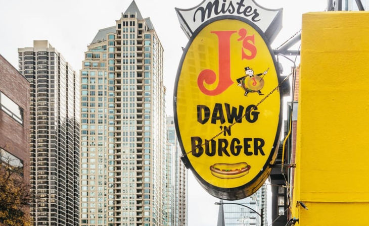 Bright yellow vintage hot dog and burger sign hanging from a city storefront with downtown high-rise buildings in the background