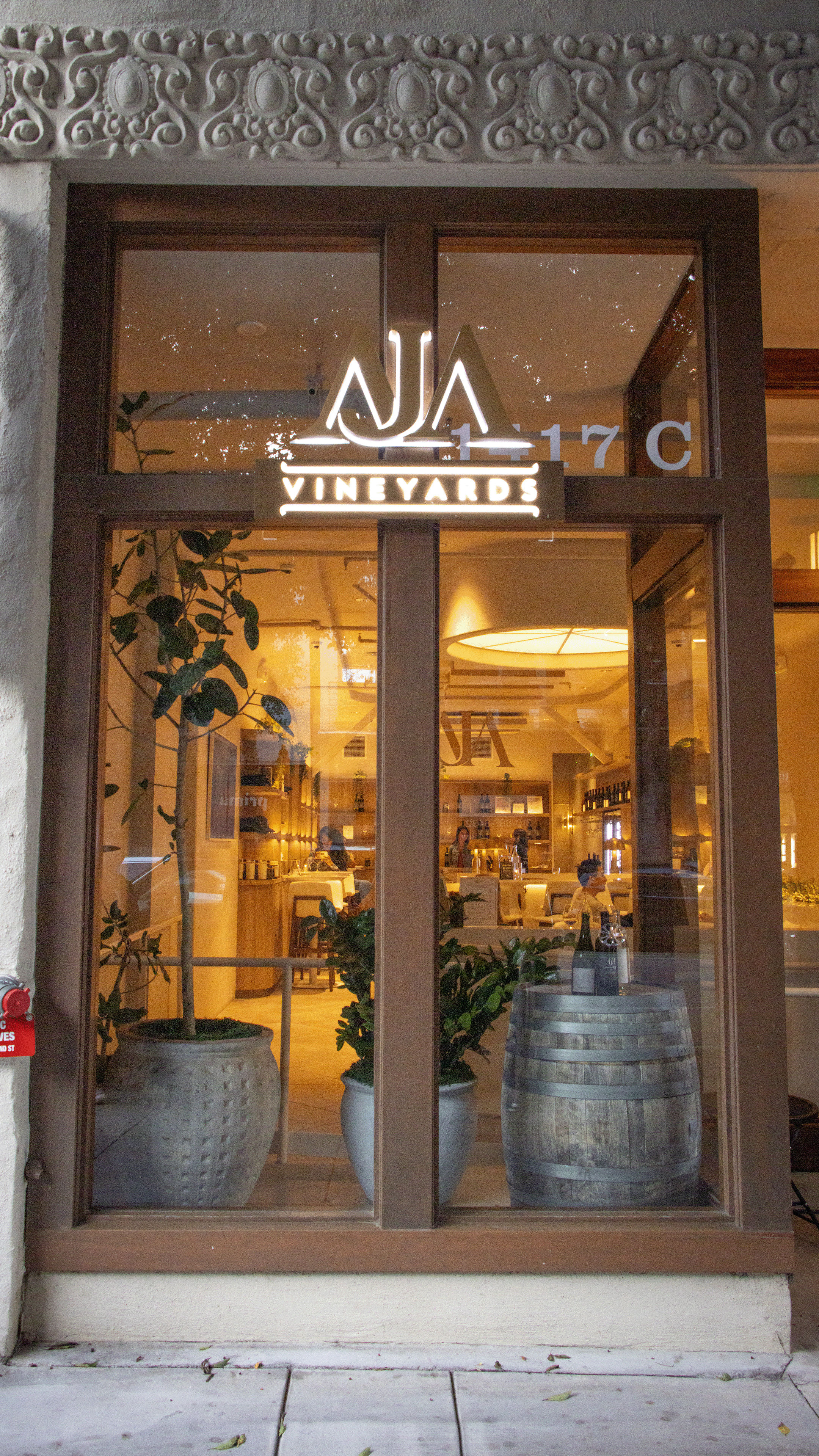 Cozy wine tasting room storefront with illuminated sign in a wooden-framed glass window, visible wooden barrel and potted plants, warm interior lighting and ornate stone lintel above the entrance.