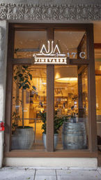 Cozy wine tasting room storefront with illuminated sign in a wooden-framed glass window, visible wooden barrel and potted plants, warm interior lighting and ornate stone lintel above the entrance.