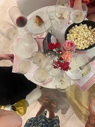 Overhead shot of a round indoor table at a casual gathering with multiple wine glasses, a bowl of popcorn, empty plates, a small pink and red rose bouquet, and guests' feet in sandals.