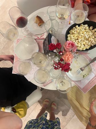 Overhead shot of a round indoor table at a casual gathering with multiple wine glasses, a bowl of popcorn, empty plates, a small pink and red rose bouquet, and guests' feet in sandals.