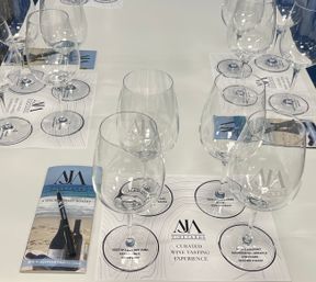 Curated wine-tasting setup with multiple empty stemmed glasses arranged on printed tasting mats, plus a brochure and pen on a white table.