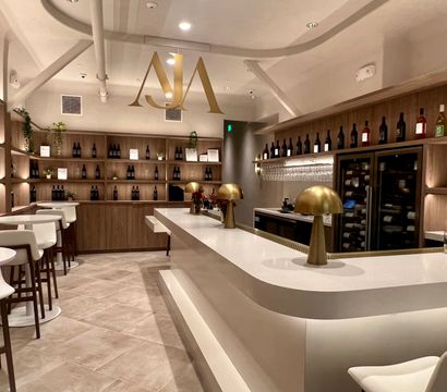 Inviting modern wine bar interior with curved white bar, brass mushroom lamps, white barstools, and wooden shelves lined with wine bottles and warm lighting.