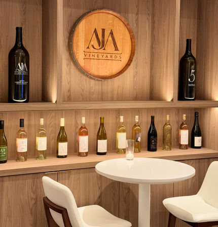 Cozy modern wine tasting nook with backlit wooden shelves of assorted white, rosé and red bottles, a decorative wooden barrel plaque, a small round white table and two sleek white chairs.